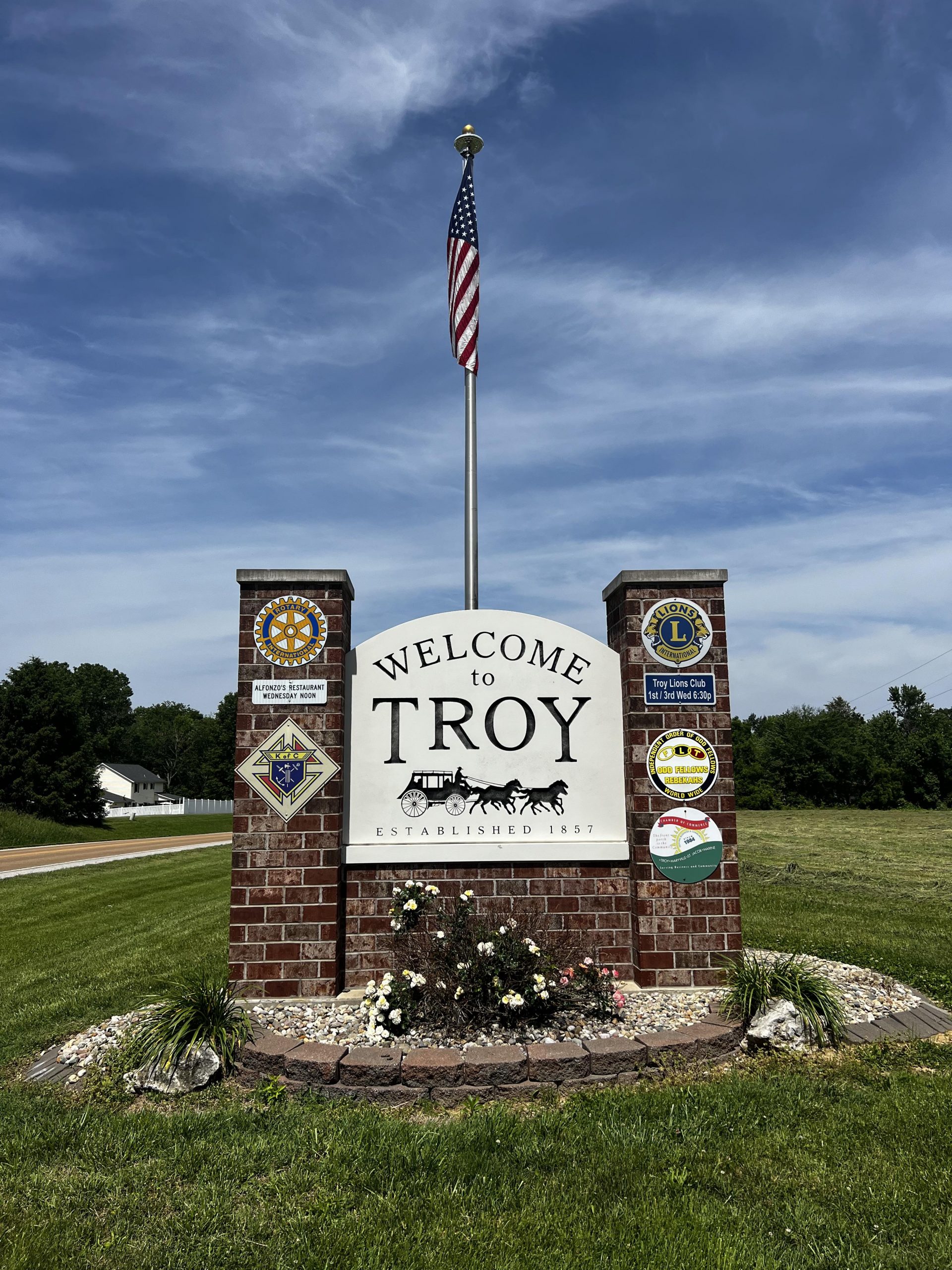 Brick and mortar sign with the words Welcome to Troy on a white background and the American Flag on a pole