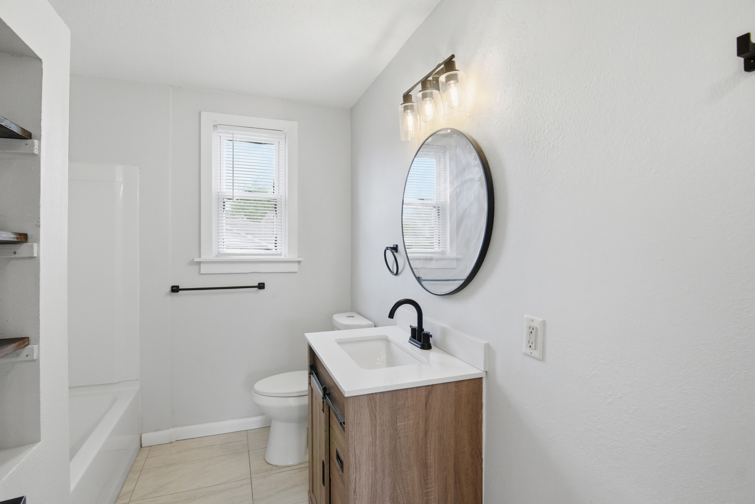 Renovated full bathroom with rustic wood-toned vanity, matte black fixtures, round mirror, modern three-light sconce, tiled floor, window with blinds, and built-in shelving at 1929 Beckwith St. Madison.