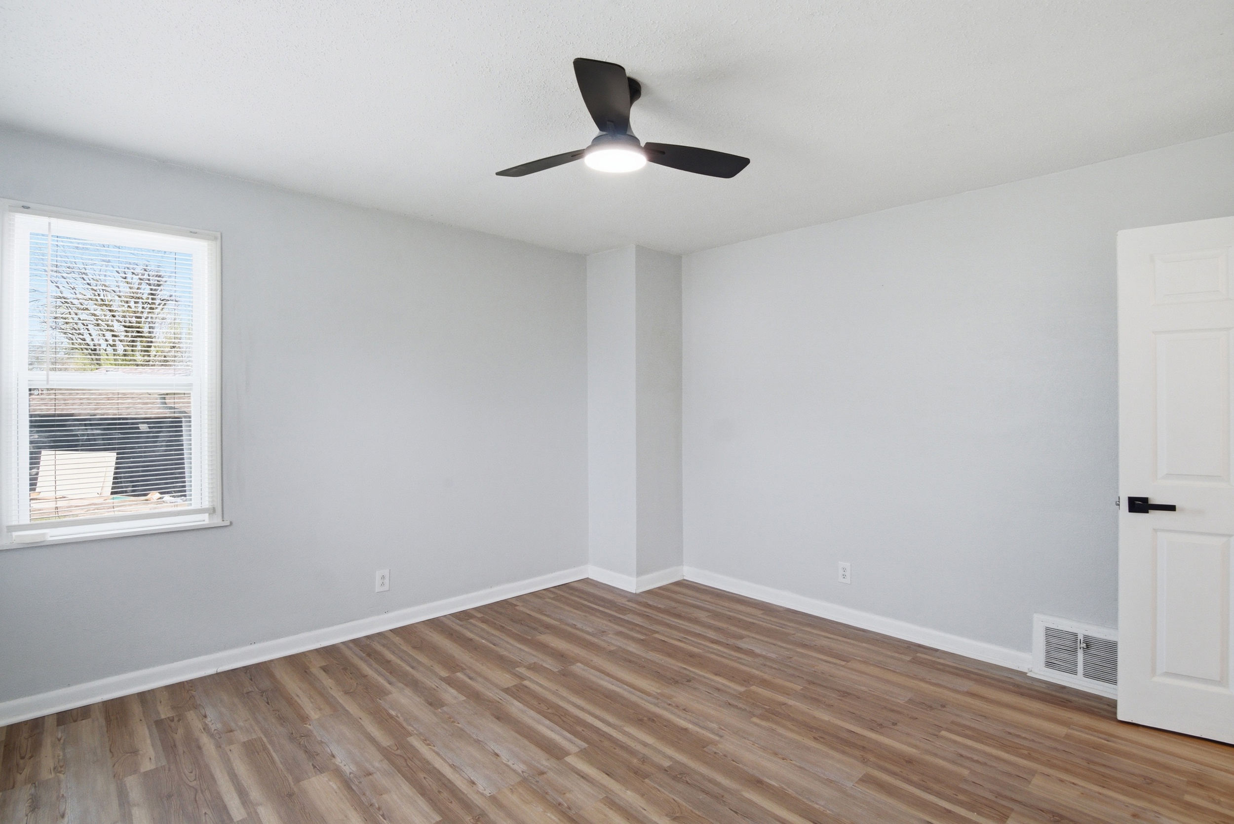 Grey walls, wood-looking- laminate flooring, black ceiling fan, and window with blinds in primary bedroom at 1929 Beckwith St. Madison.