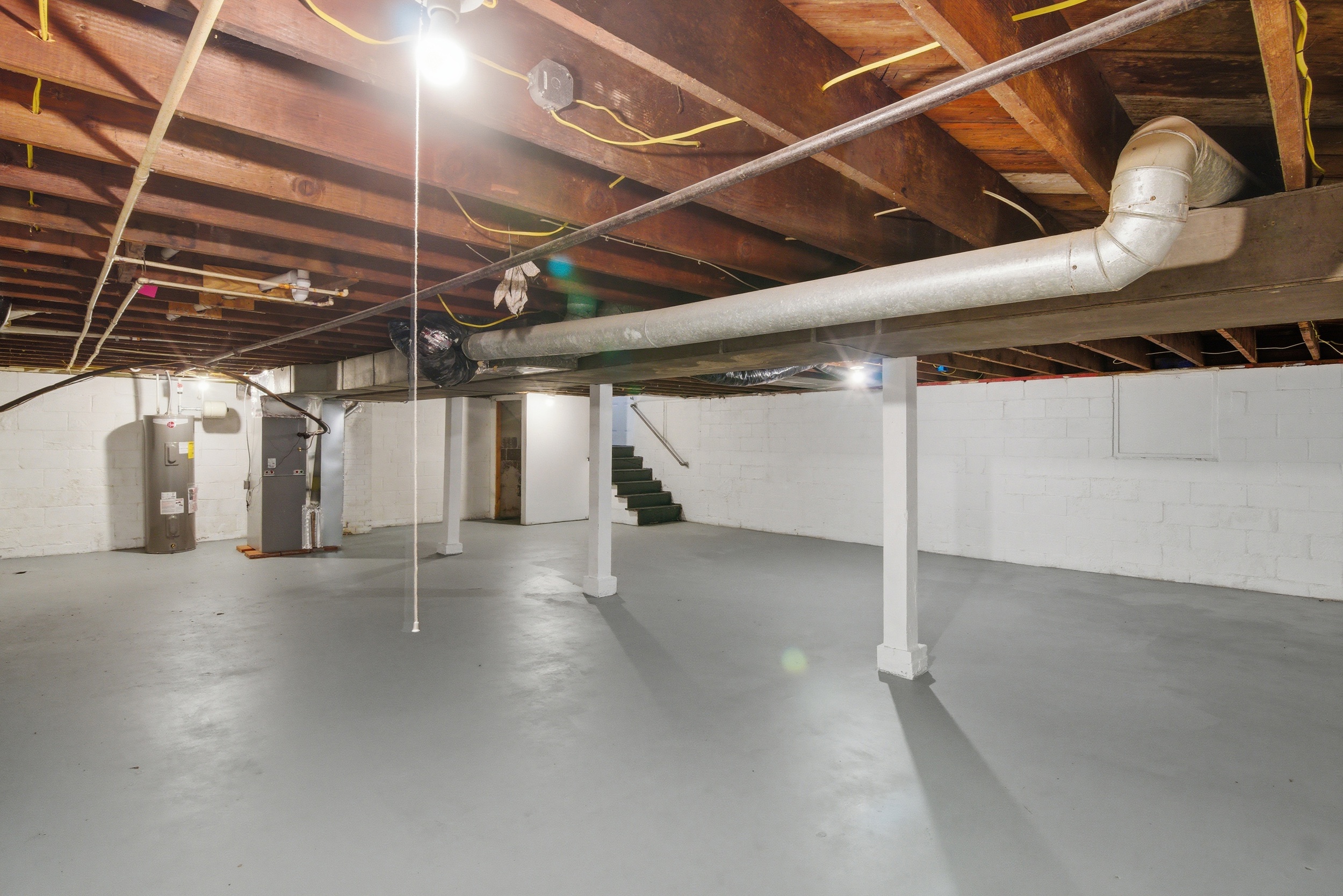 painted basement mechanical area featuring a hot water heater, furnace unit painted concrete walls, and staircase with dark treads at 1929 Beckwith St. Madison.
