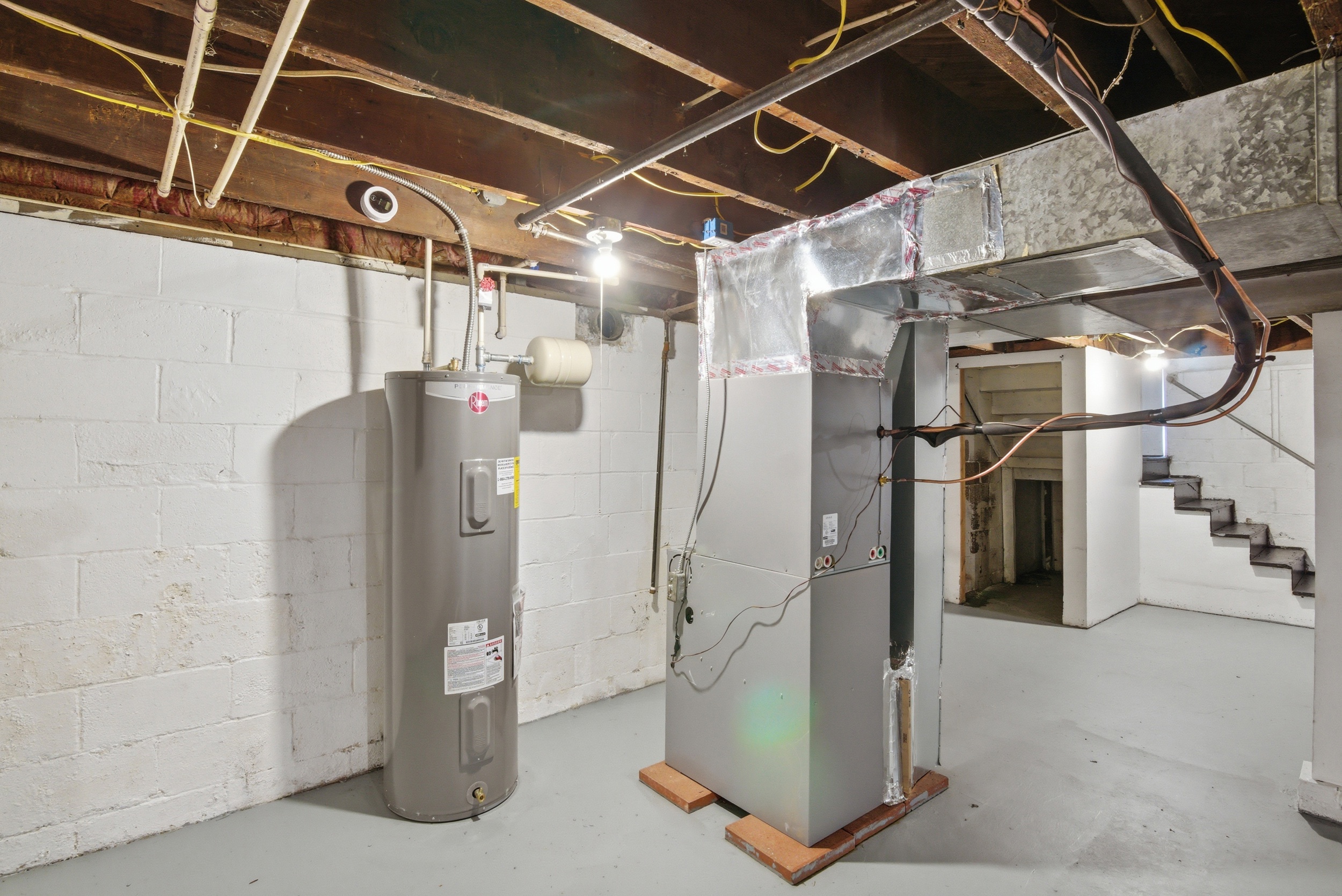 Clean, painted basement mechanical area featuring a Rheem hot water heater, grey furnace unit with silver ductwork, painted concrete walls, and staircase with dark treads at 1929 Beckwith St. Madison.