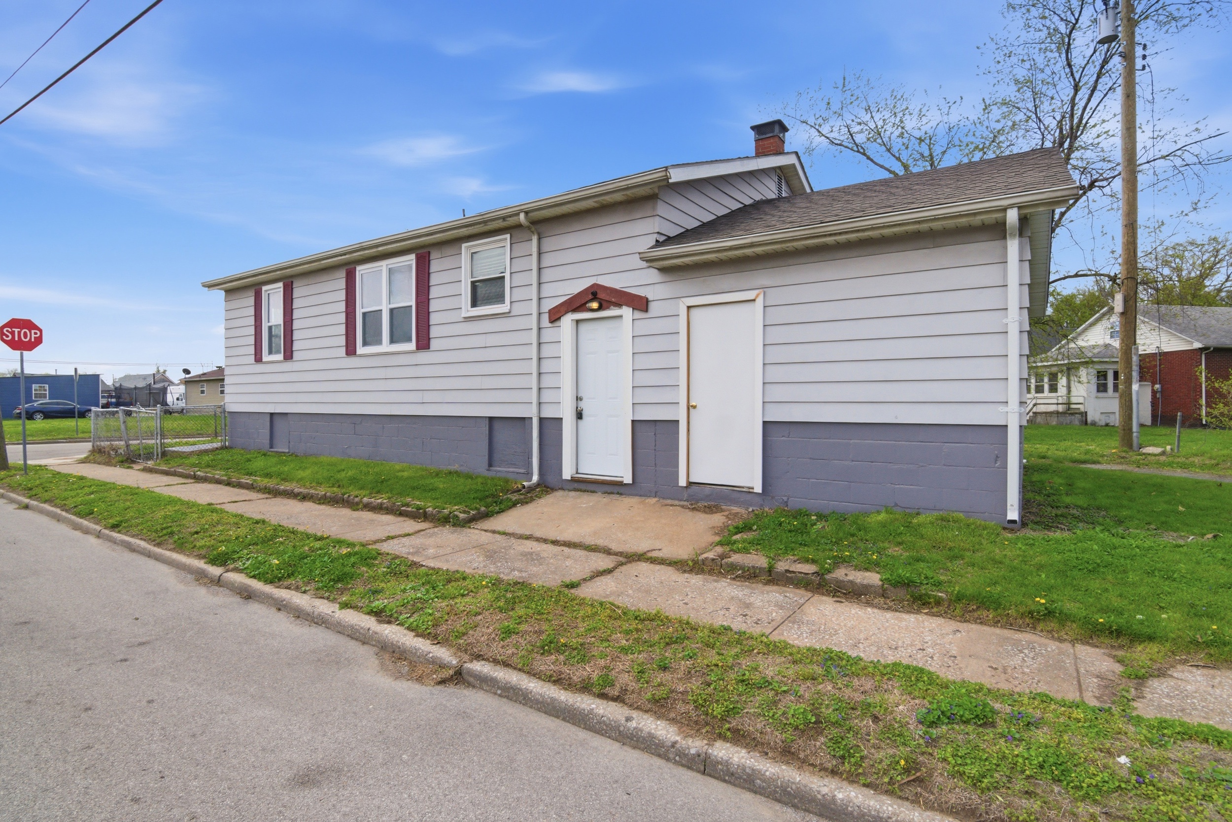 Side angled of the exterior side of the property featuring grey siding, dark maroon shutters, two white doors, small concrete pad and concrete sidewalk at 1929 Beckwith St. Madison.