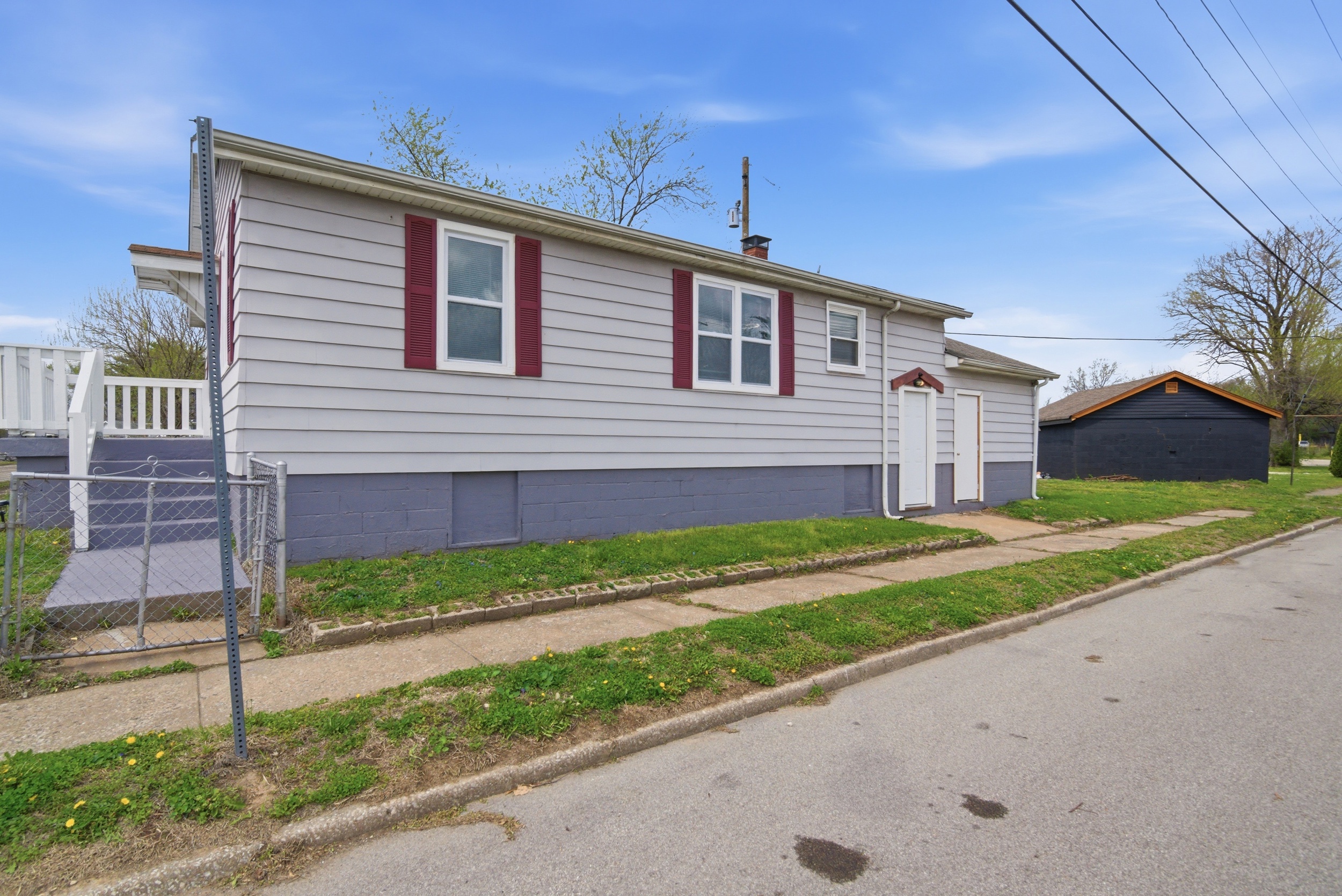 Exterior view of the Street Side of the house featuring grey Aluminum siding, dark maroon shutters, white windows, a gray/blue painted foundation, and two white doors leading to a small concrete pad at 1929 Beckwith St. Madison.