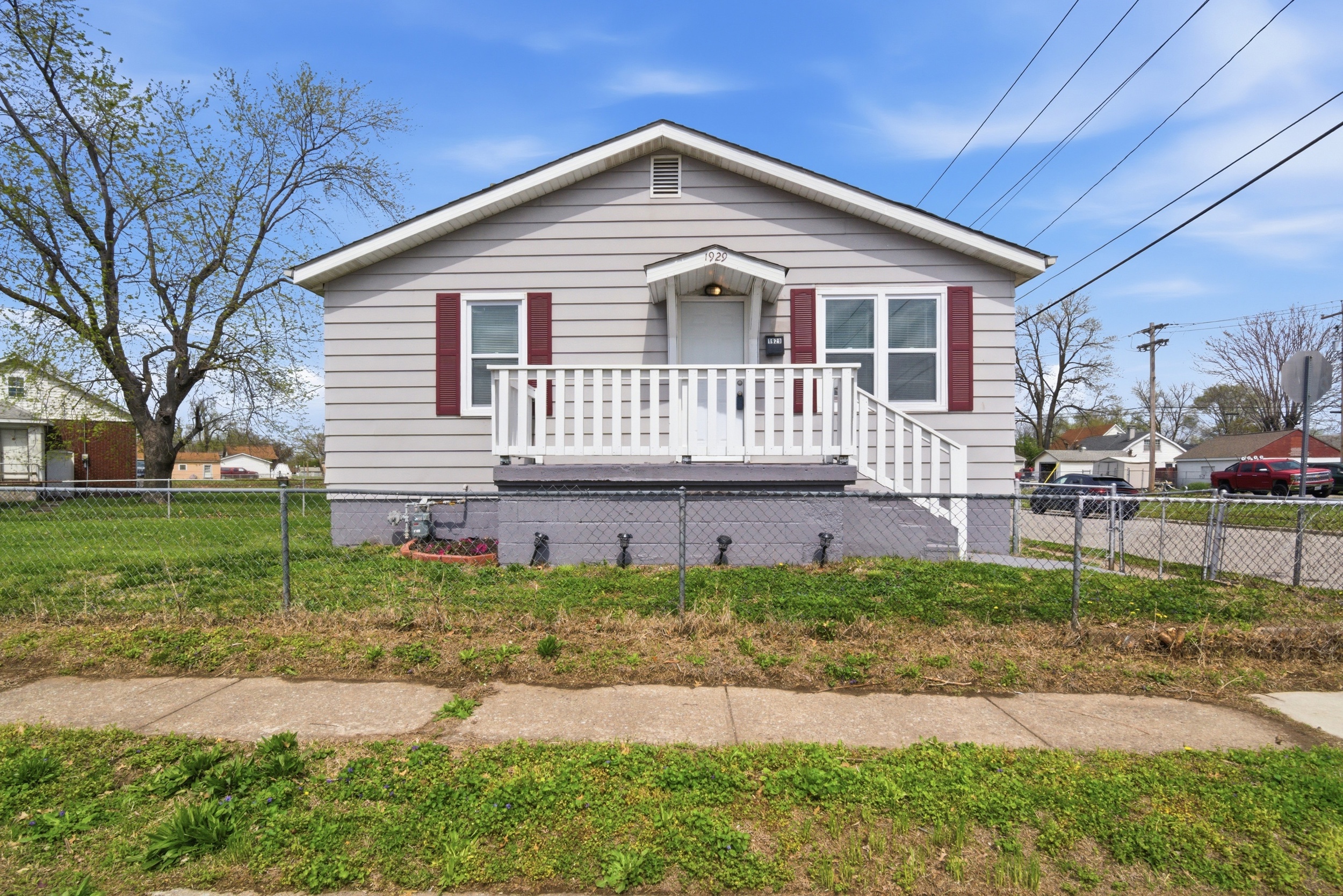 Exterior front view of a grey-sided house with maroon shutters, a white front porch with railings, and a fenced front yard with a concrete sidewalk at 1929 Beckwith St, Madison. The scene is under a clear blue sky.